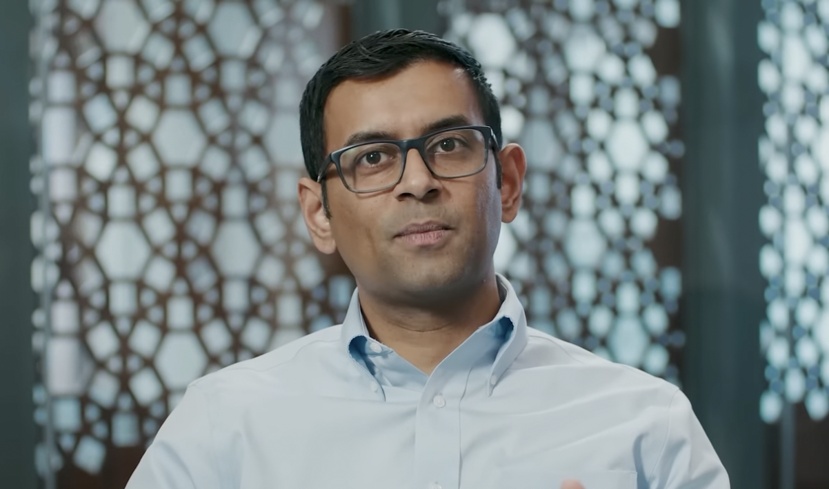 Professor Arvind Narayanan is sitting and discussing his work at CITP. He is wearing a light blue shirt and glasses. There is a patterned wall behind him.